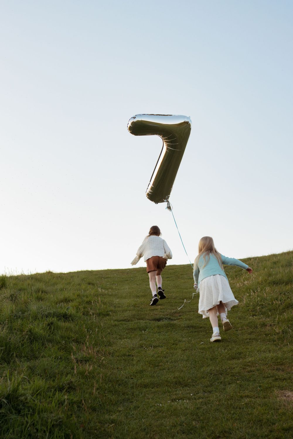Two children running up a green hill holding a large silver number 7 balloon against a clear blue sky.