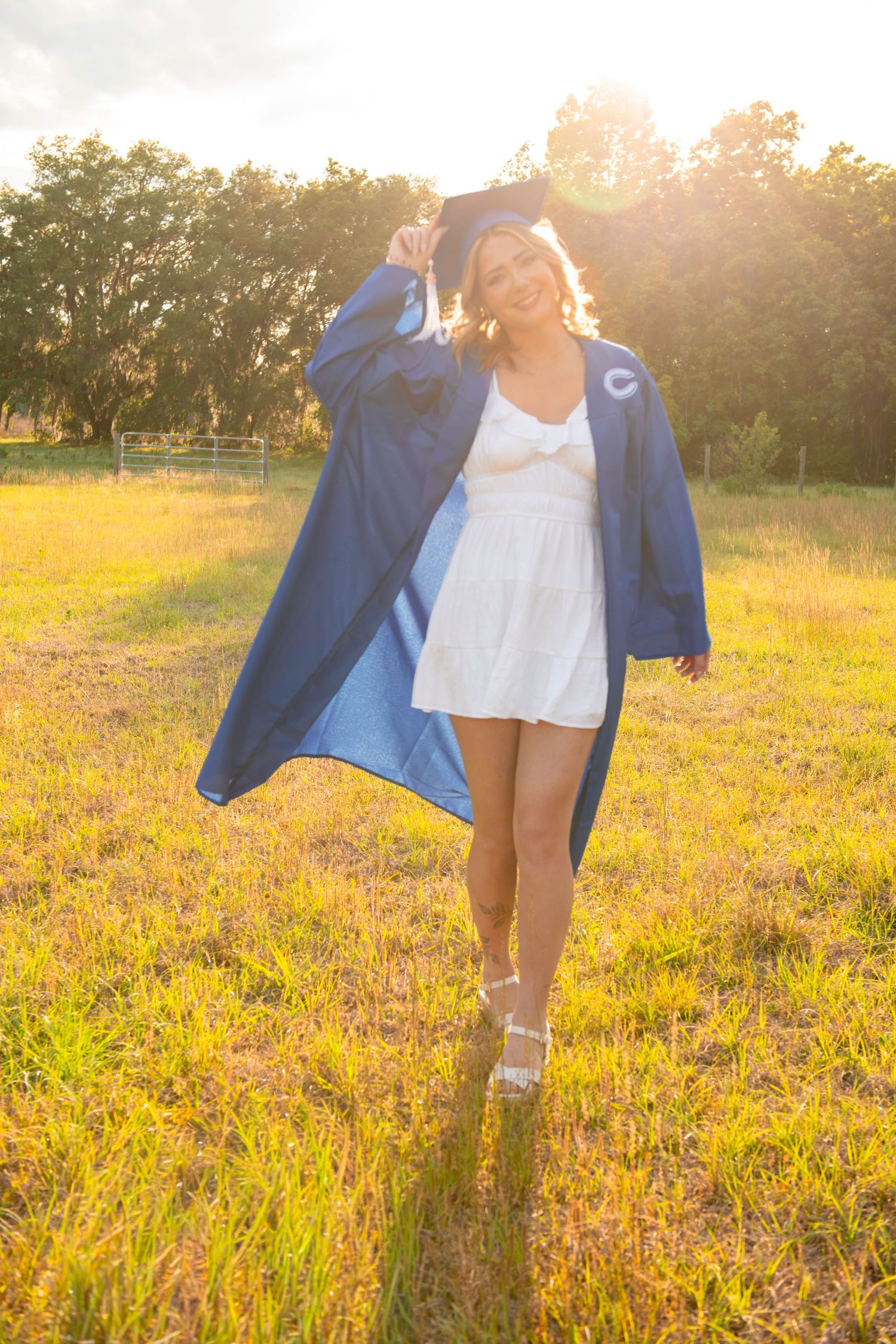 Young woman in blue graduation cap and gown over white dress celebrating outdoors in sunlit field.