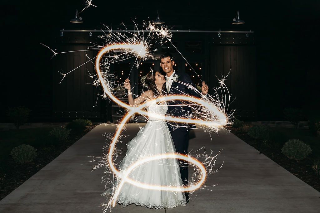 Bride and groom pose with swirling sparklers creating light trails at night outside a wedding venue.
