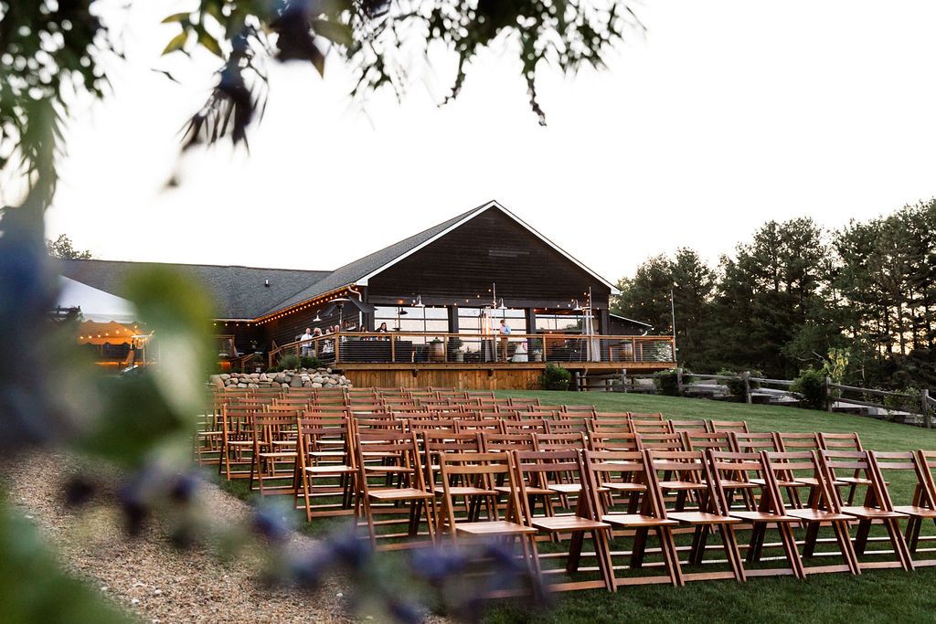 Outdoor wedding ceremony setup with rows of wooden folding chairs on a lawn facing a rustic barn venue surrounded by trees.