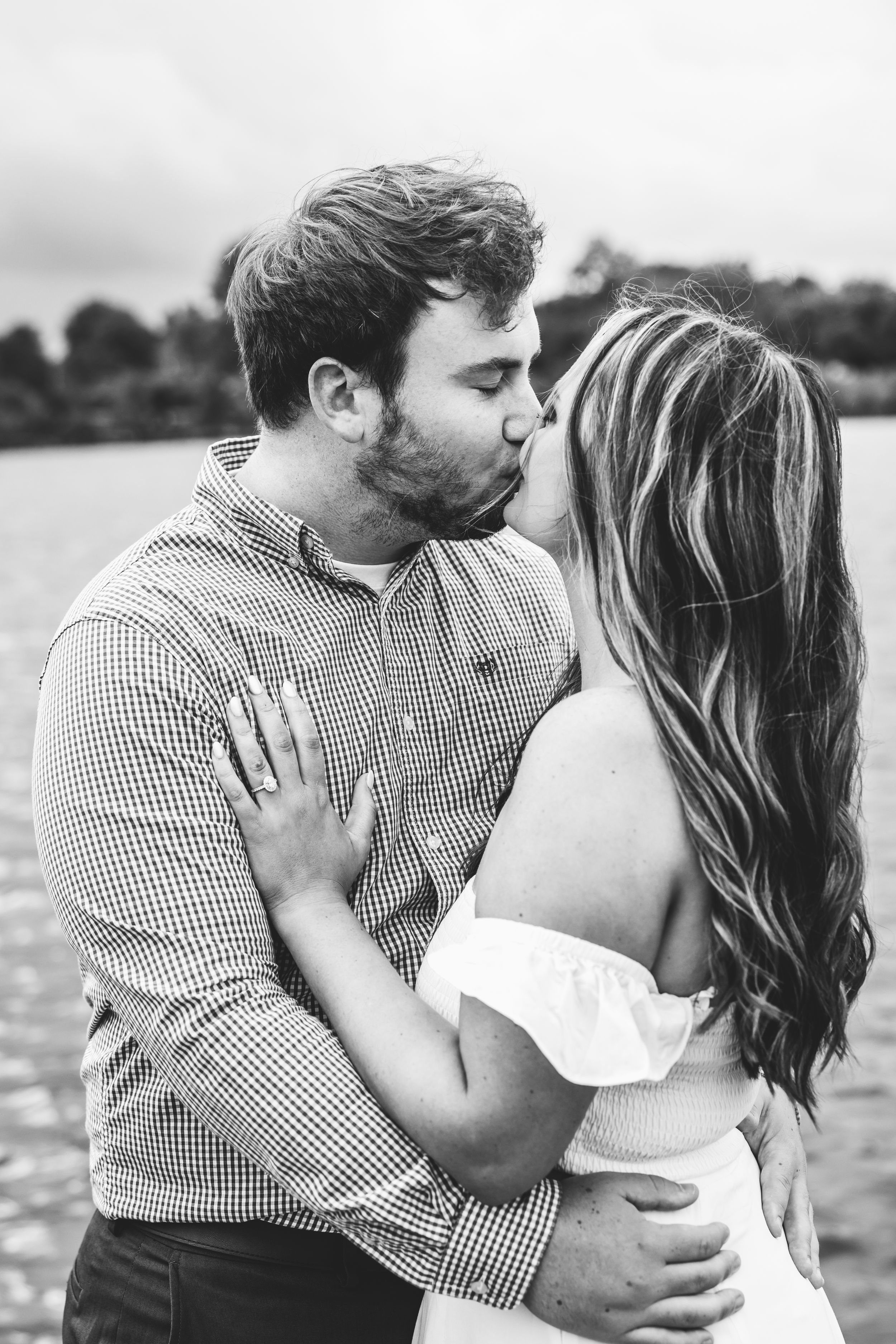 Black and white photo of a couple sharing a romantic kiss outdoors near a lake, woman wearing white dress.