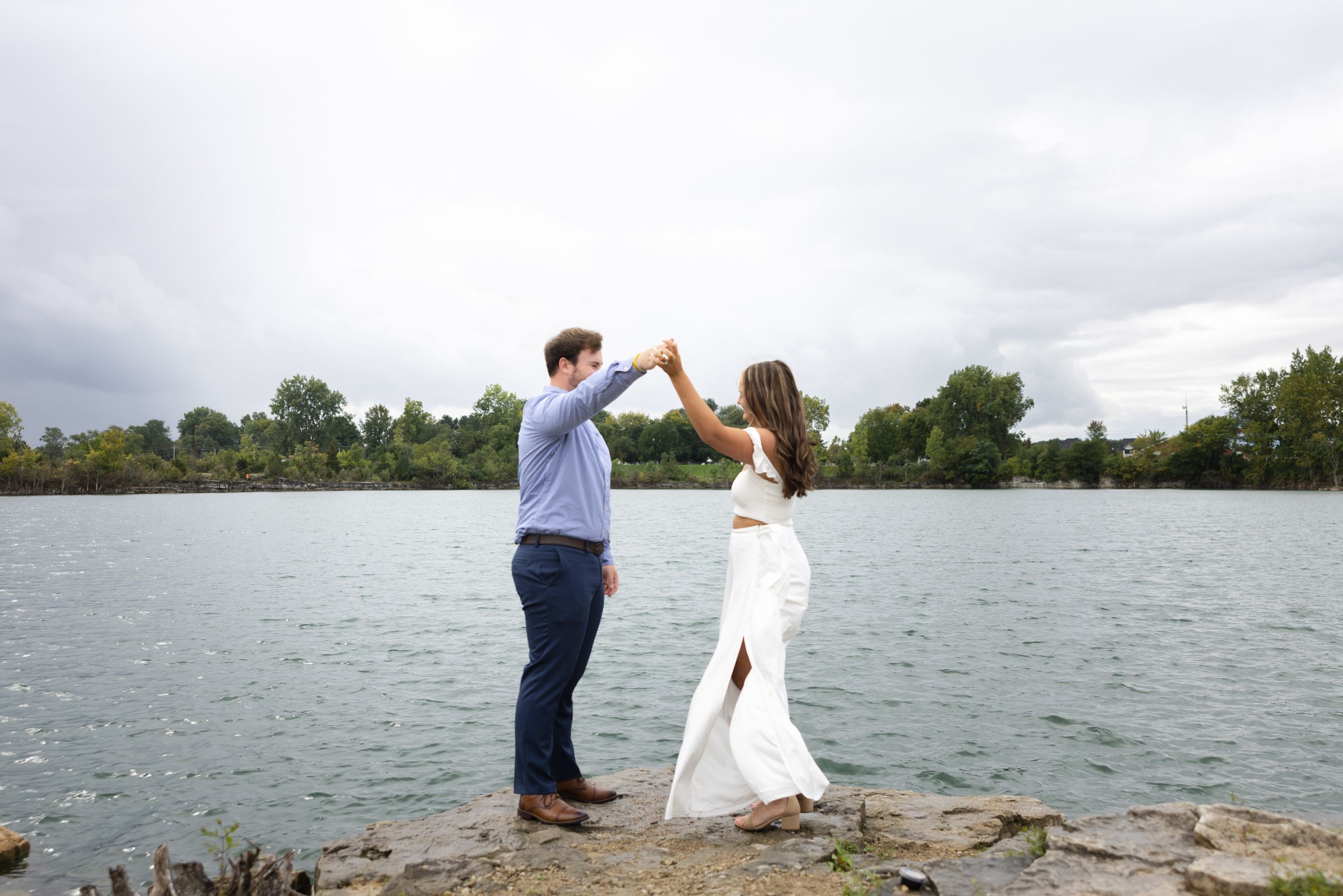 Couple dancing hand in hand on rocky shore beside a calm lake outdoors.