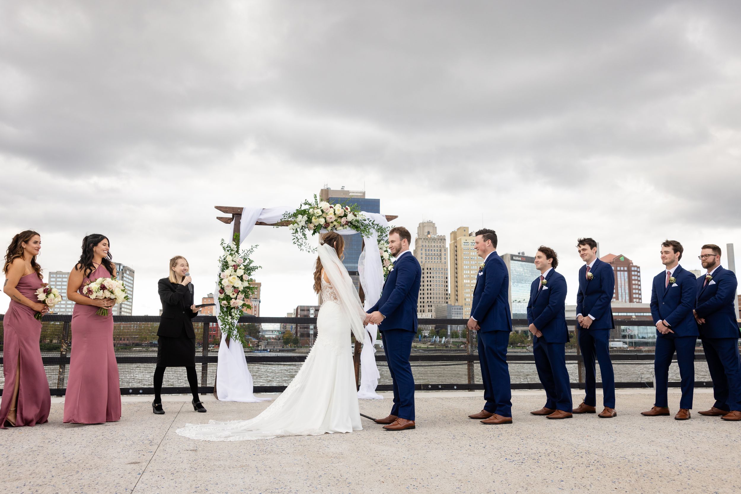 Outdoor rooftop wedding ceremony with bridal party in navy suits and mauve dresses, city skyline backdrop.