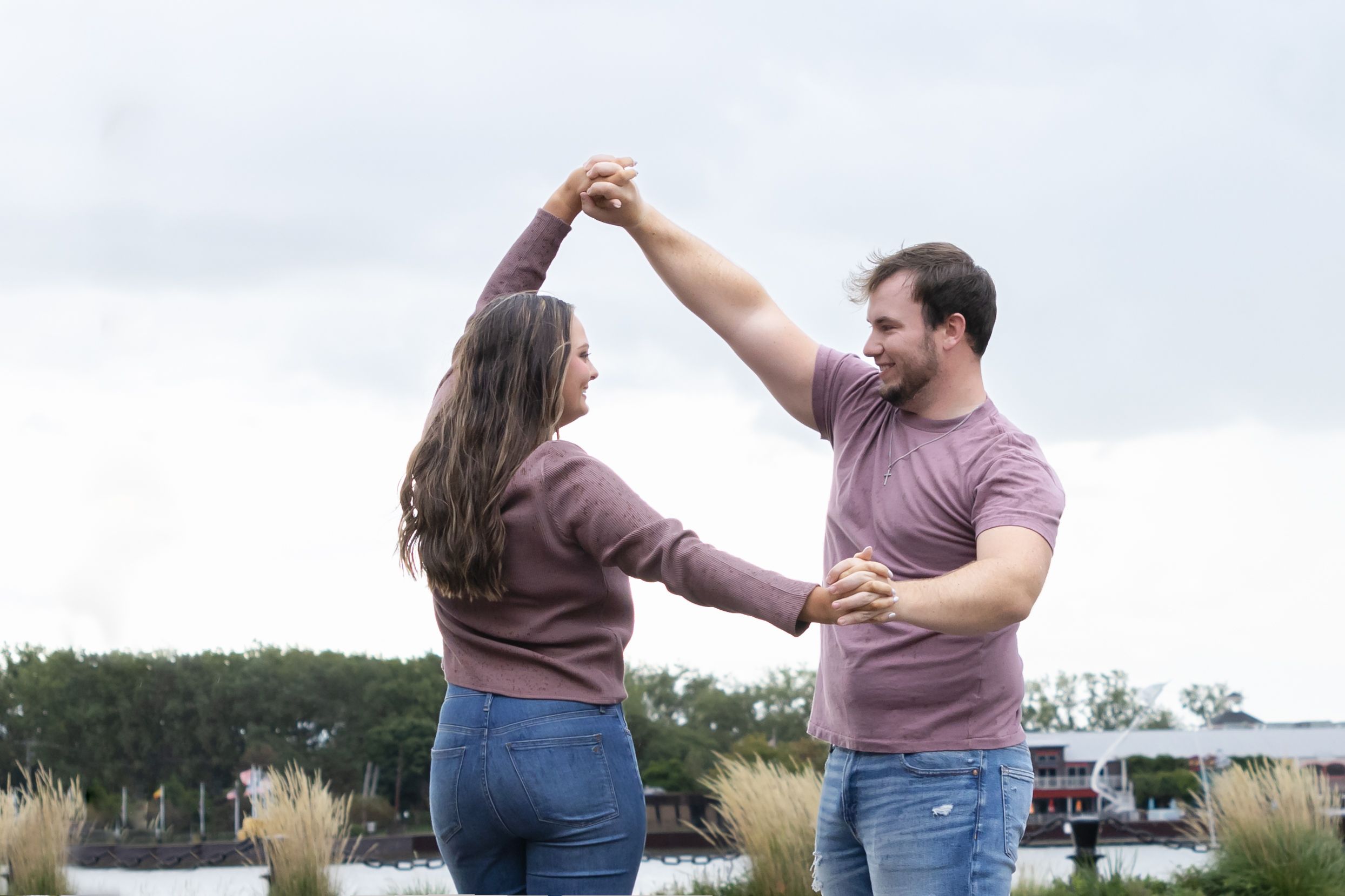 Happy couple dancing outdoors in matching mauve tops and jeans with scenic waterfront background.