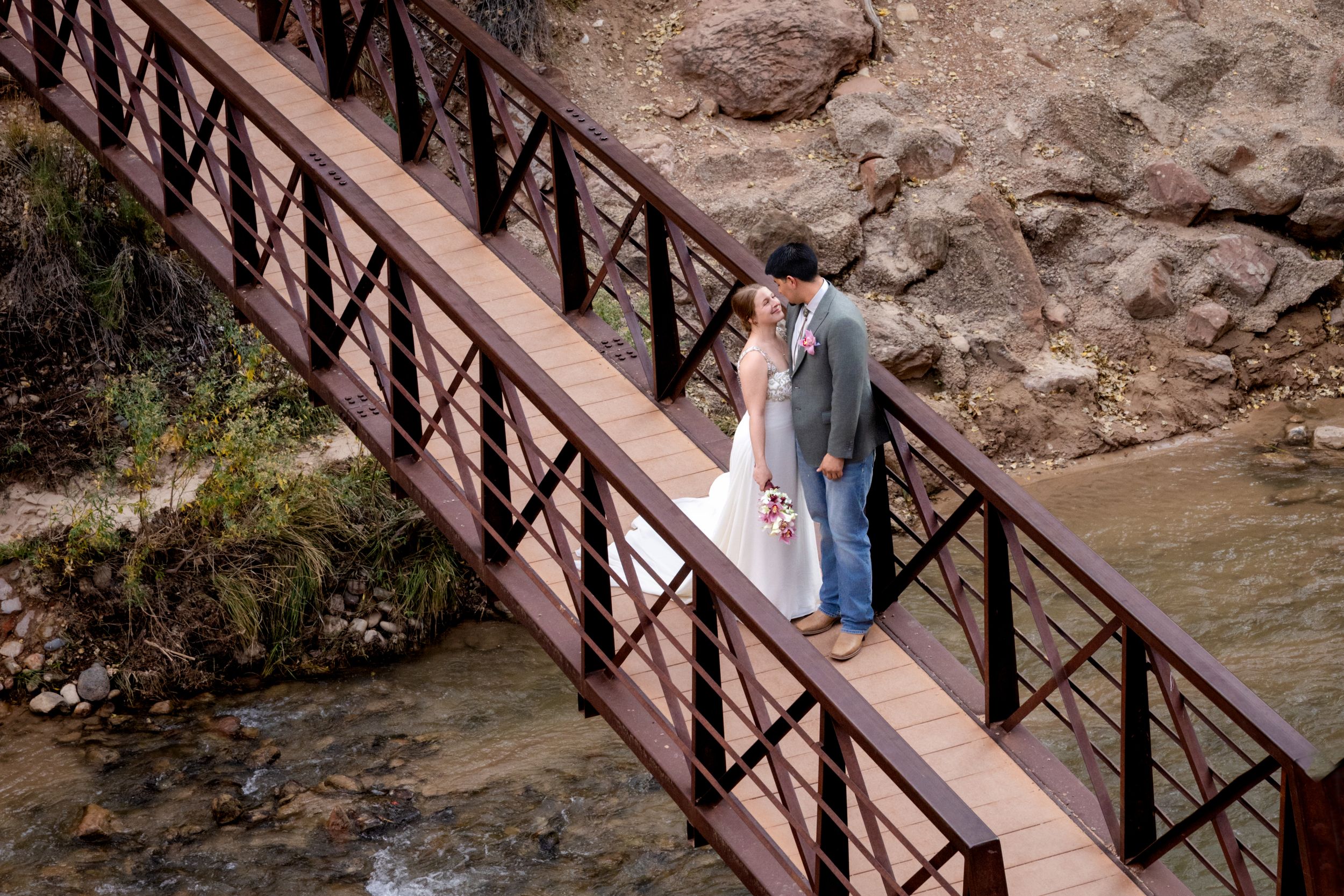 Bride and groom share a romantic kiss on a wooden bridge over a mountain stream during their wedding day.