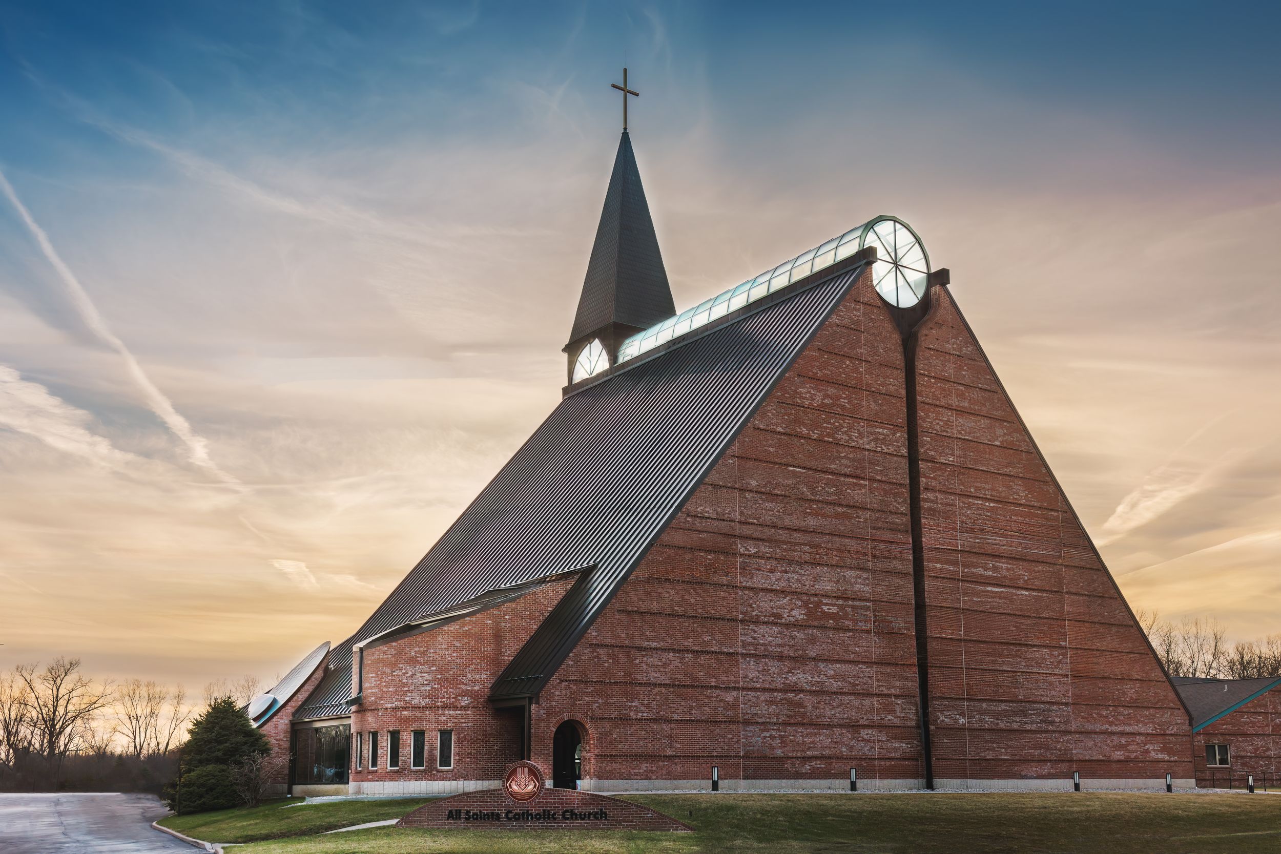 Modern triangular brick church with a cross steeple and clock under a dramatic sunset sky.