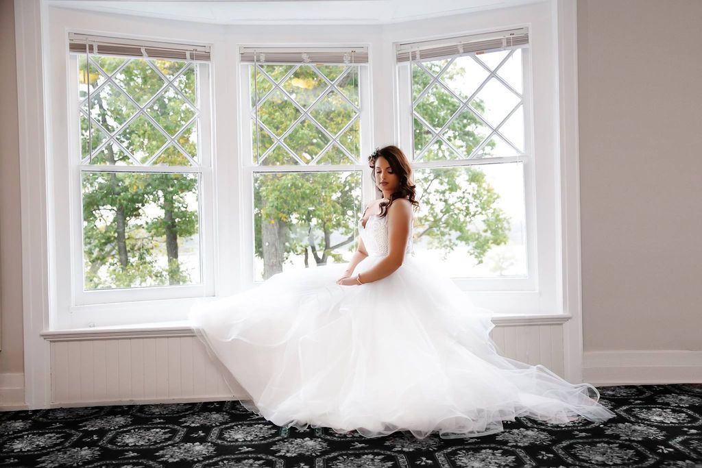 Bride in a stunning white ballgown wedding dress sitting by bay windows with natural light.