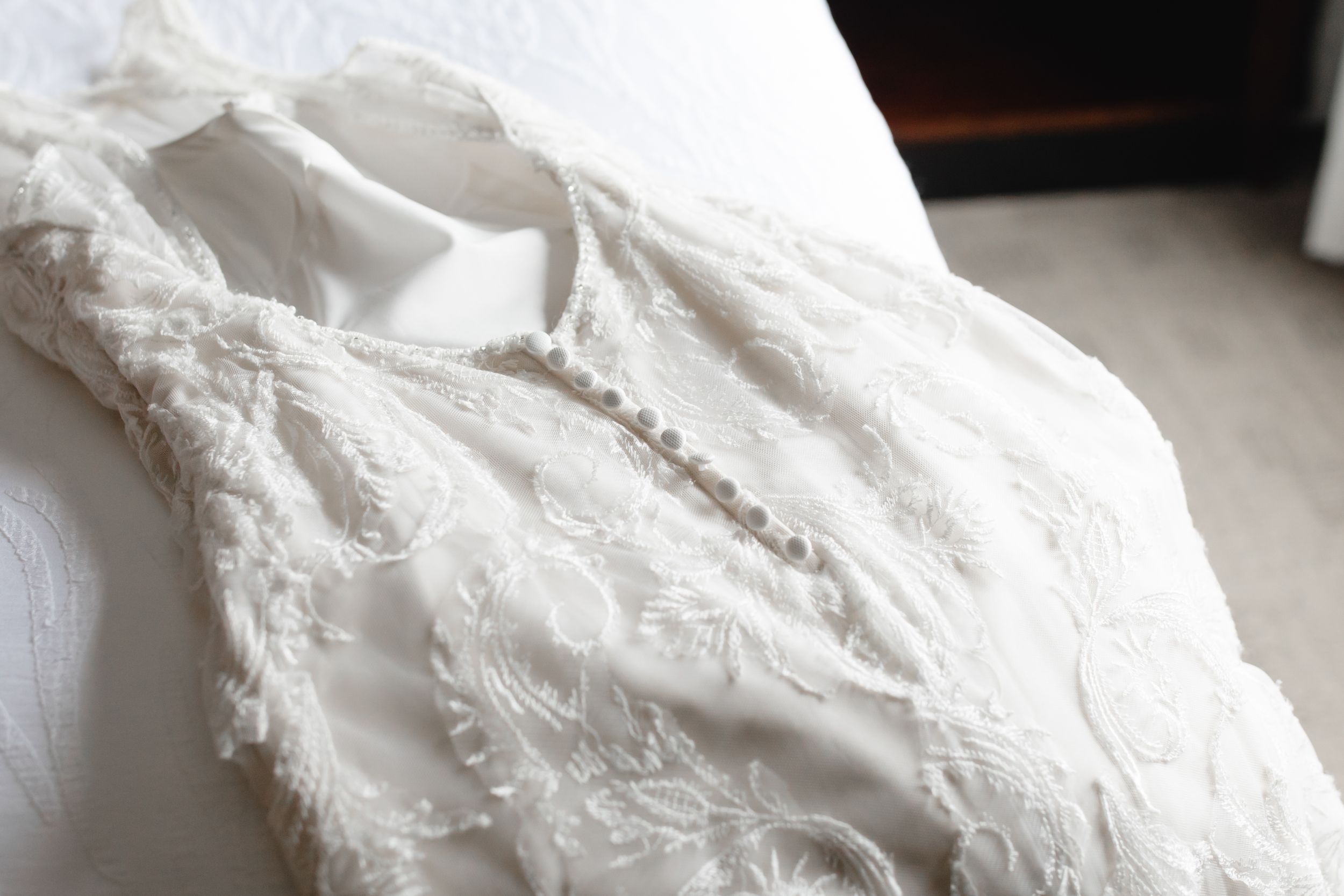 White lace wedding dress with floral embroidery and pearl button details laid flat on a white surface.