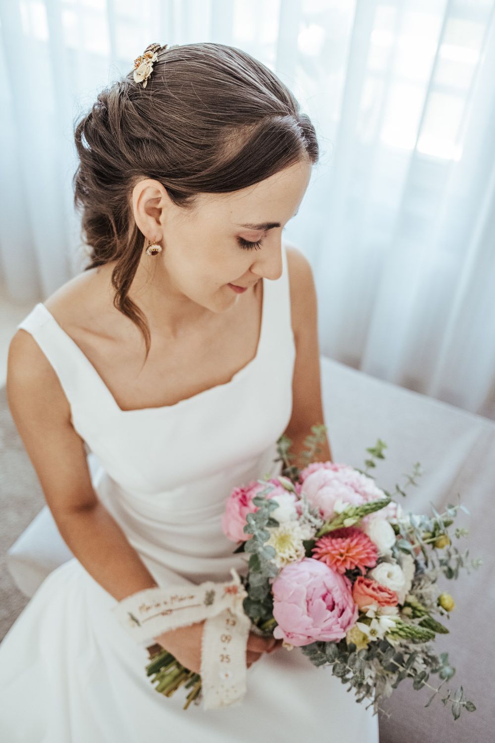 Bride in white dress holds colorful bouquet of pink peonies and roses, seated by window on wedding day.