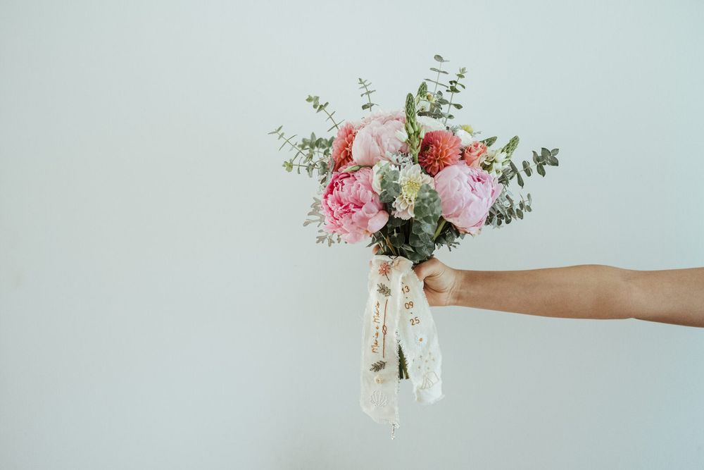 Hand holding a bridal bouquet of pink peonies, roses, and eucalyptus with a lace ribbon on white background.