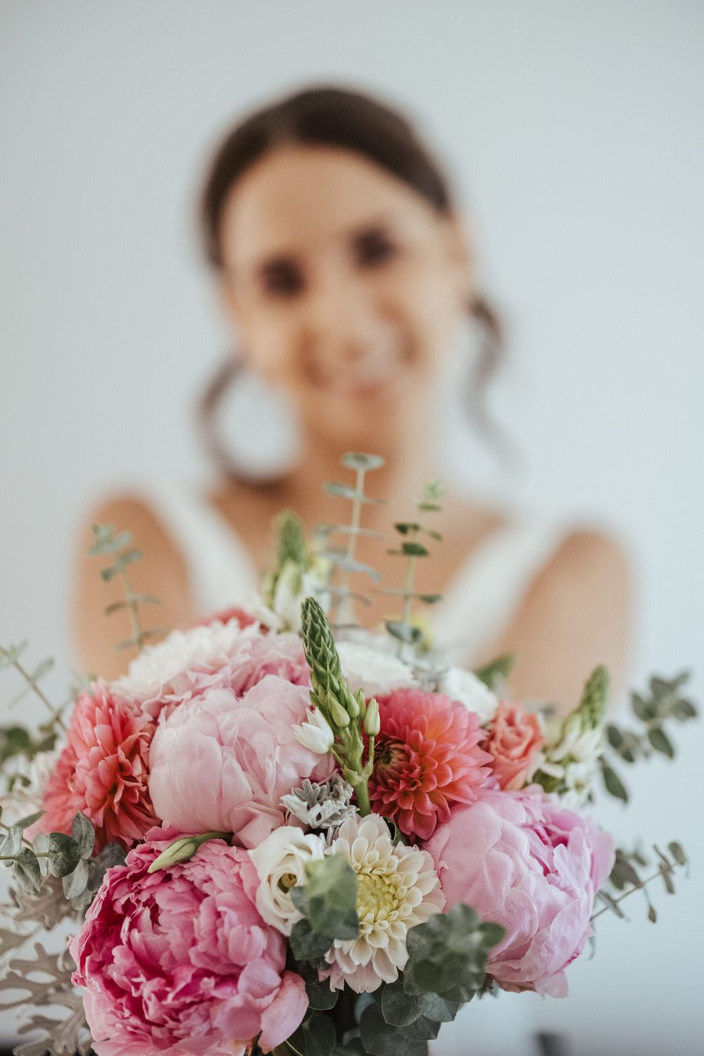Smiling bride in white dress holding a beautiful bouquet of pink peonies, dahlias, and eucalyptus.