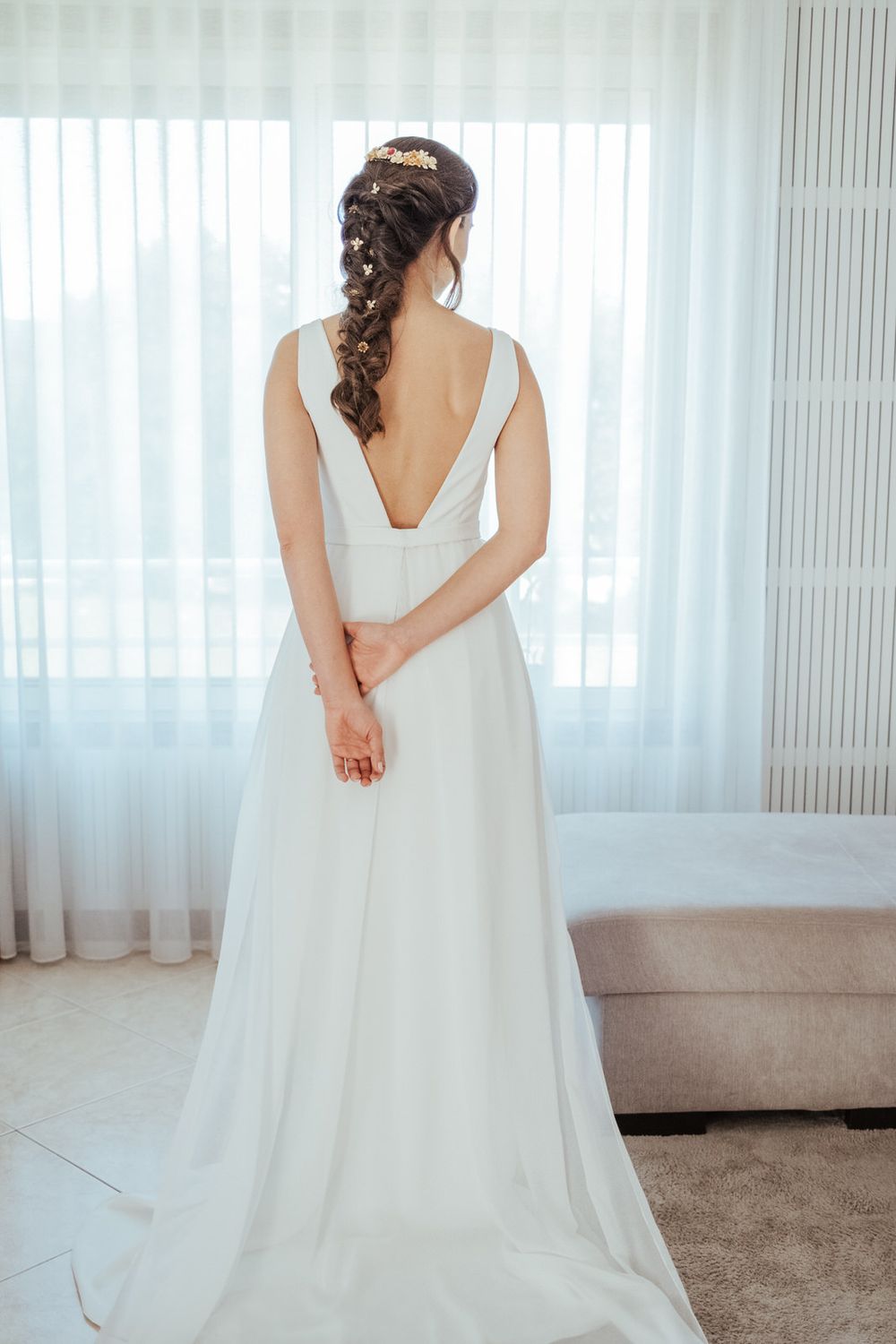 Bride in elegant white backless wedding gown with braided updo hairstyle standing by sheer curtained window.