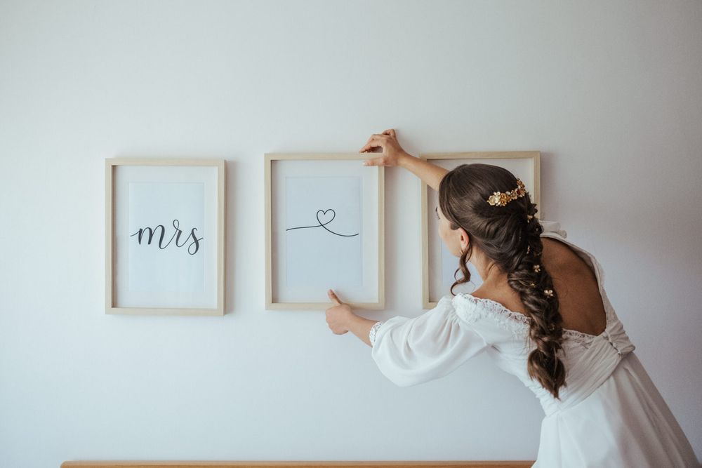 Bride in white dress hanging framed Mrs wedding sign on white wall with braided hair and floral accessory.