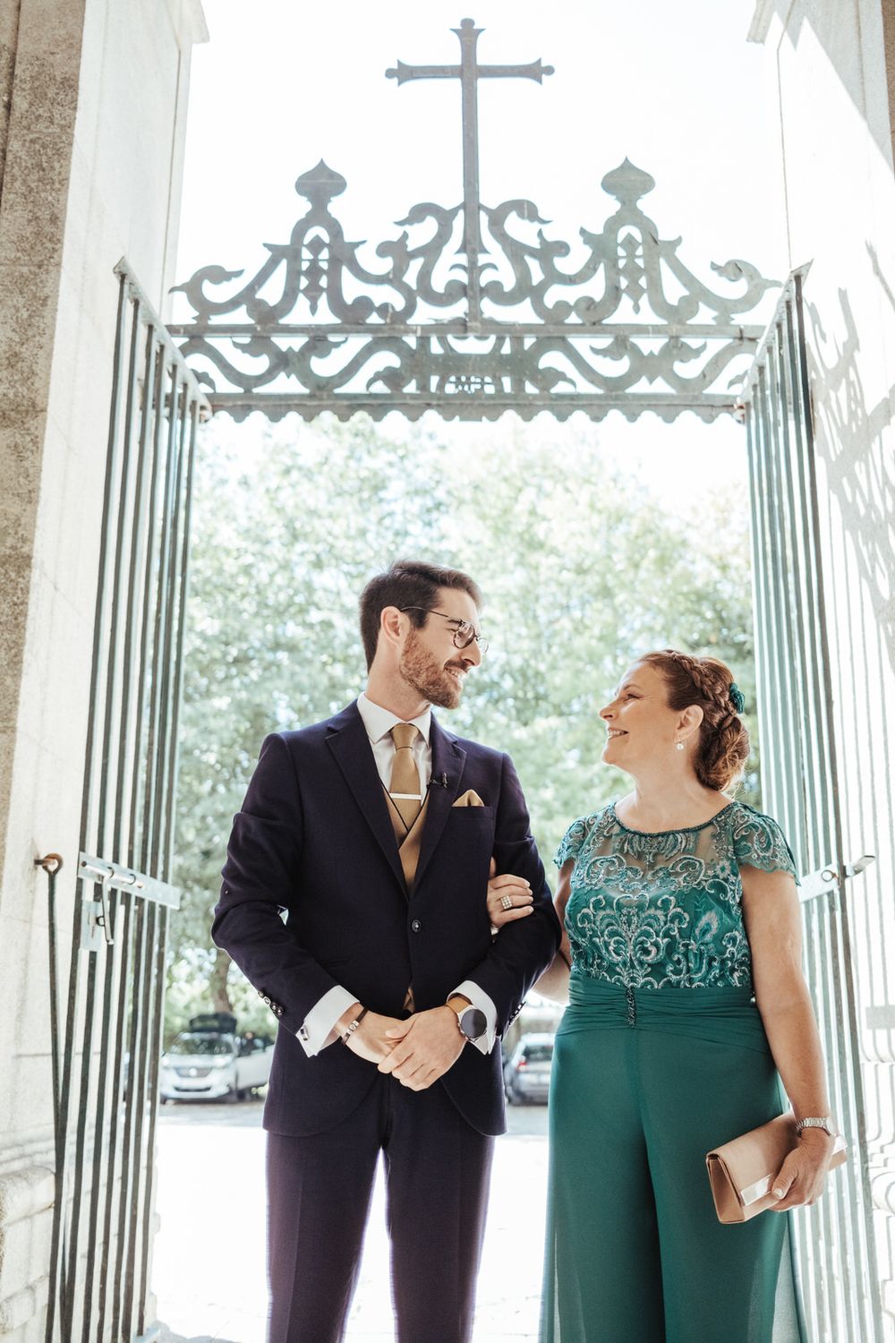 Elegant couple smiling at each other in formal attire outside ornate church gates with a cross.