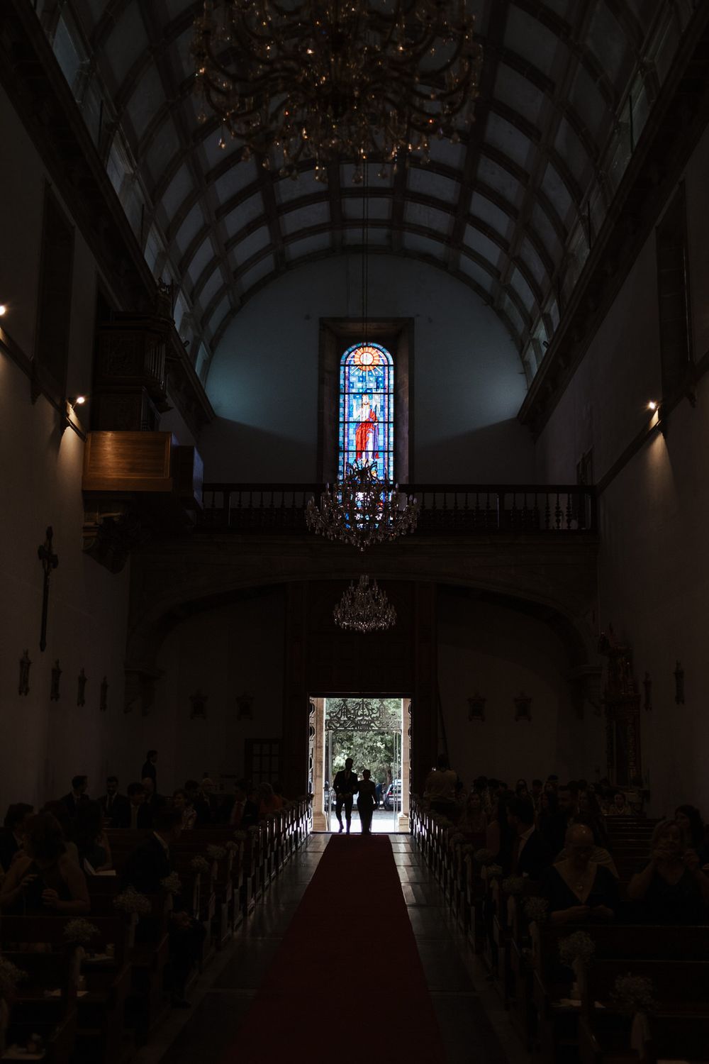 Silhouetted couple stands at church entrance with red carpet aisle, crystal chandeliers, and stained glass window above.