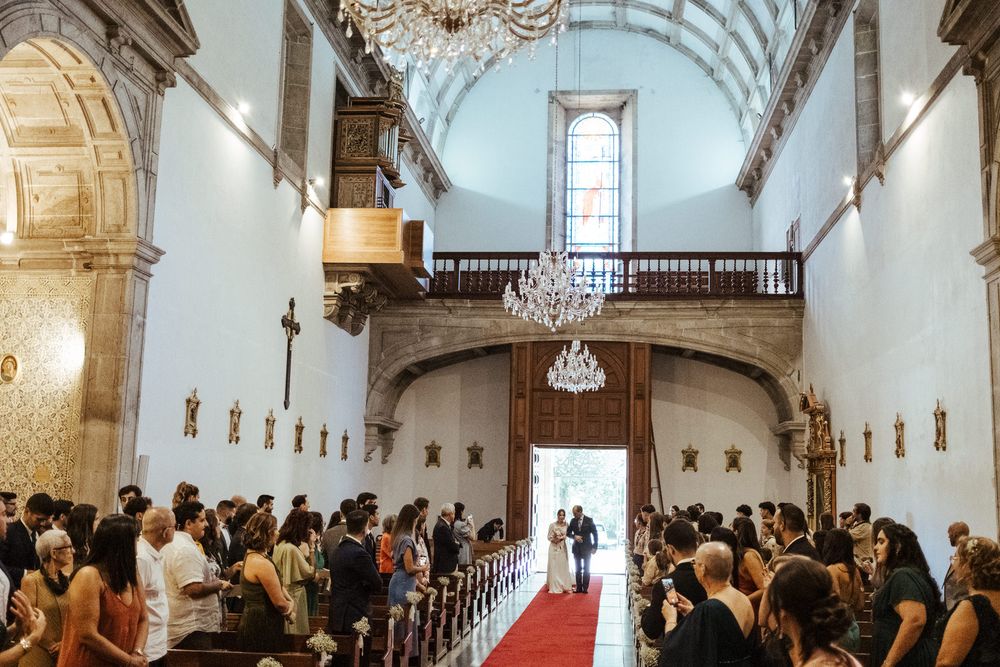 Wedding ceremony in an ornate white church with red carpet aisle, chandelier, and guests seated in wooden pews.