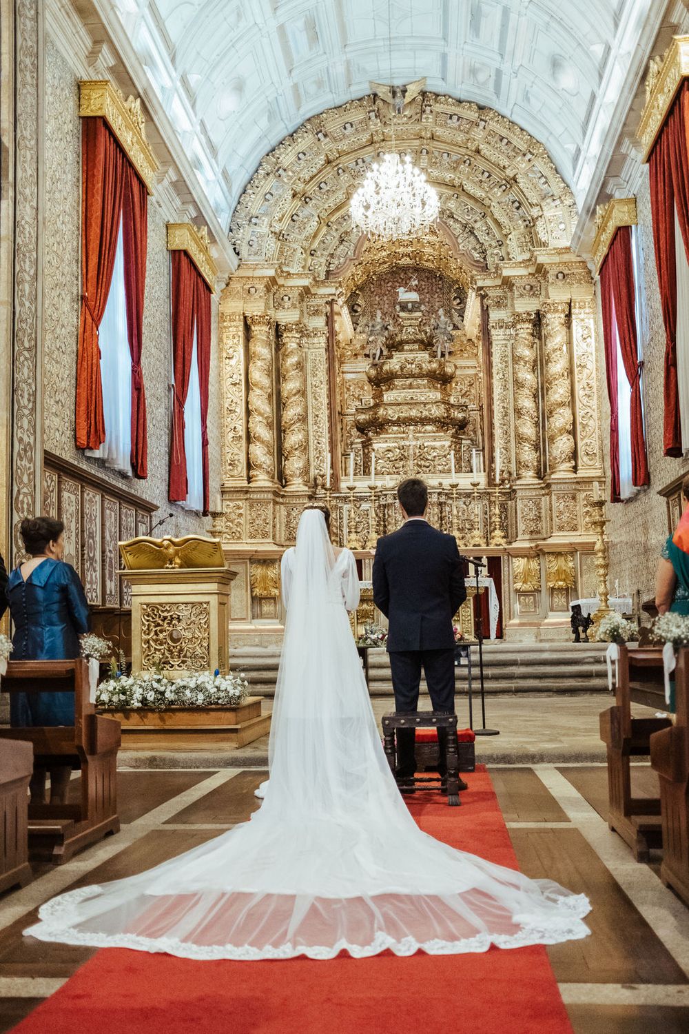 Bride in white gown with long veil stands with groom before elaborate gilded baroque church altar.