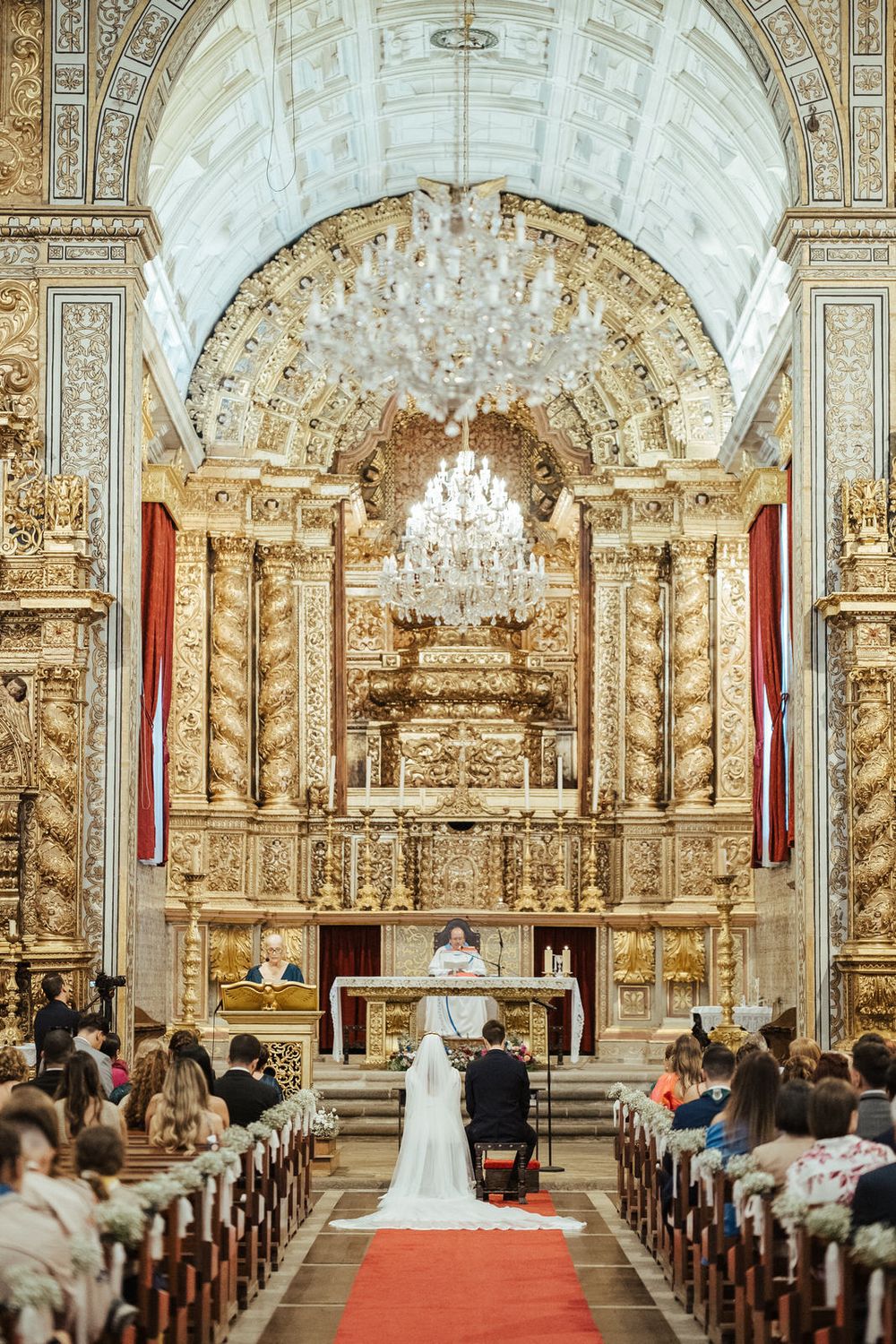 Wedding couple kneels before priest at ornate gold baroque altar with crystal chandelier above guests.