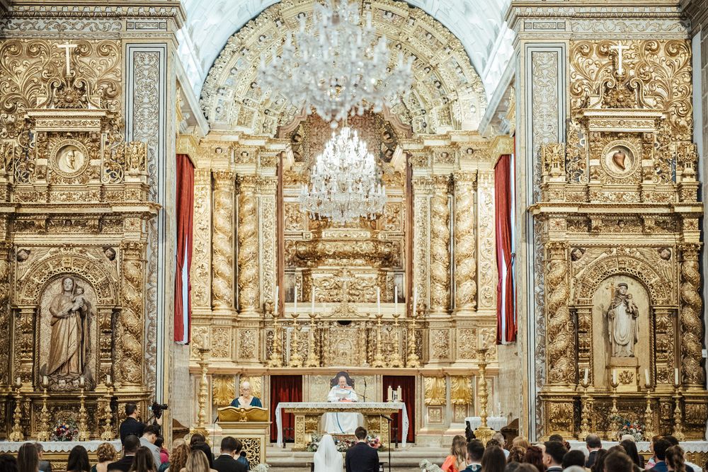 Ornate golden baroque church altar with chandelier during a wedding ceremony with seated guests.