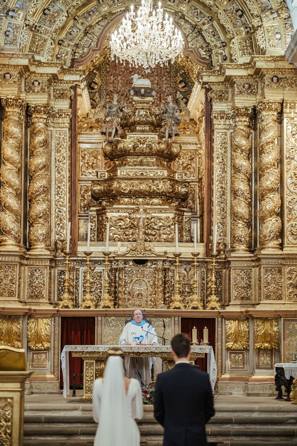 Bride and groom stand before ornate golden baroque altar during Catholic wedding ceremony in grand cathedral.