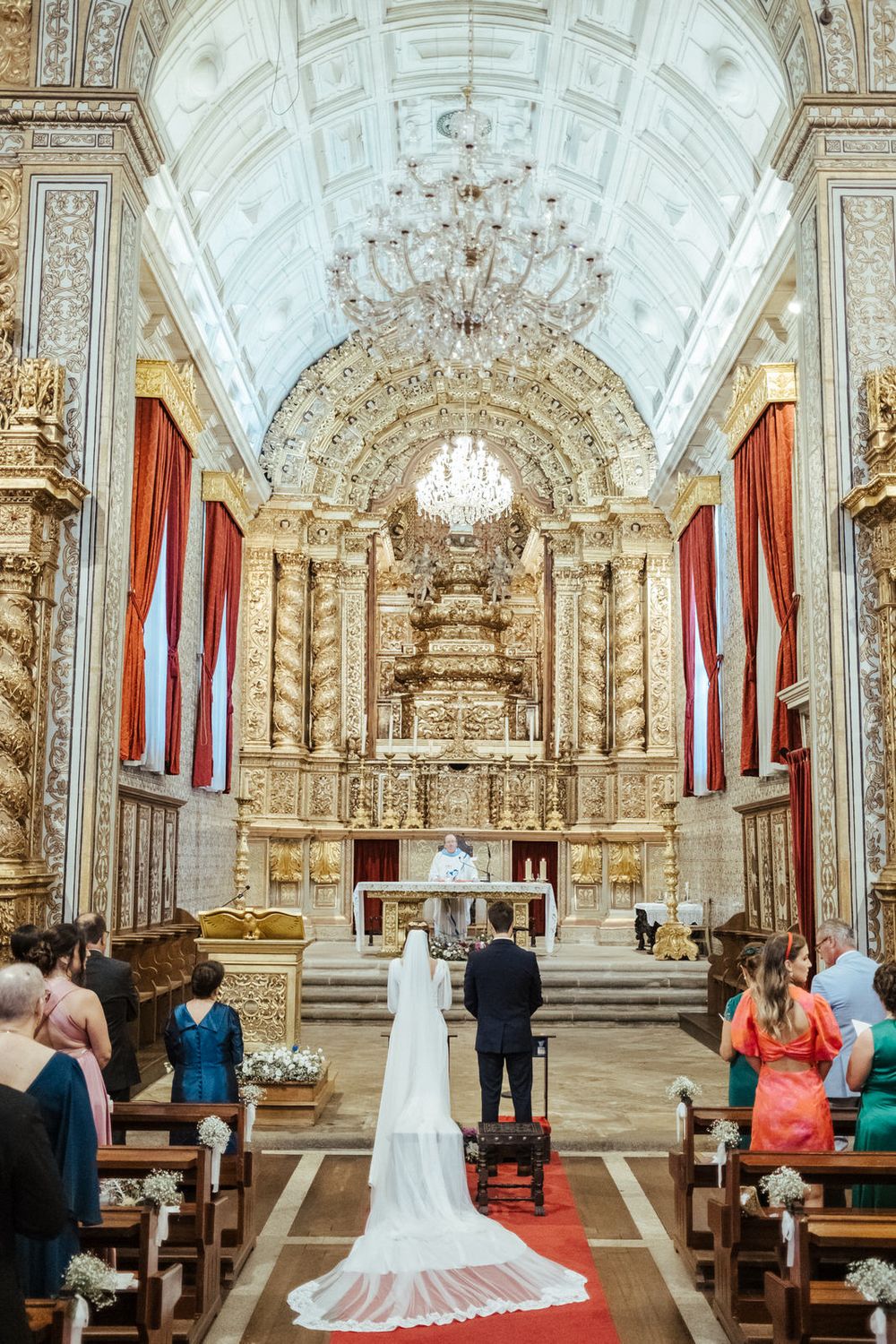 Bride and groom stand at ornate golden altar during wedding ceremony in historic baroque church with chandelier.