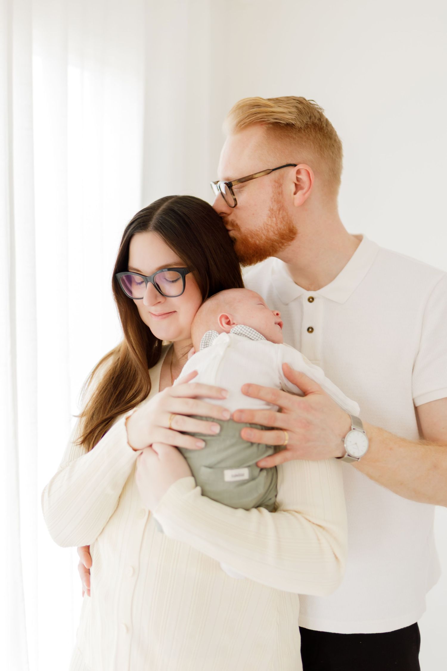 Nyblivna föräldrar håller ömt sitt nyfödda barn, med pappa som kysser mammas huvud i en ljus, minimalistisk miljö.