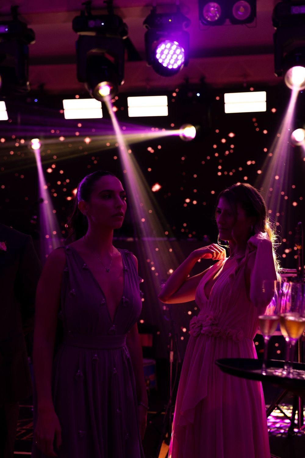 Two elegant women in flowing gowns at a glamorous event under colorful purple stage lights.
