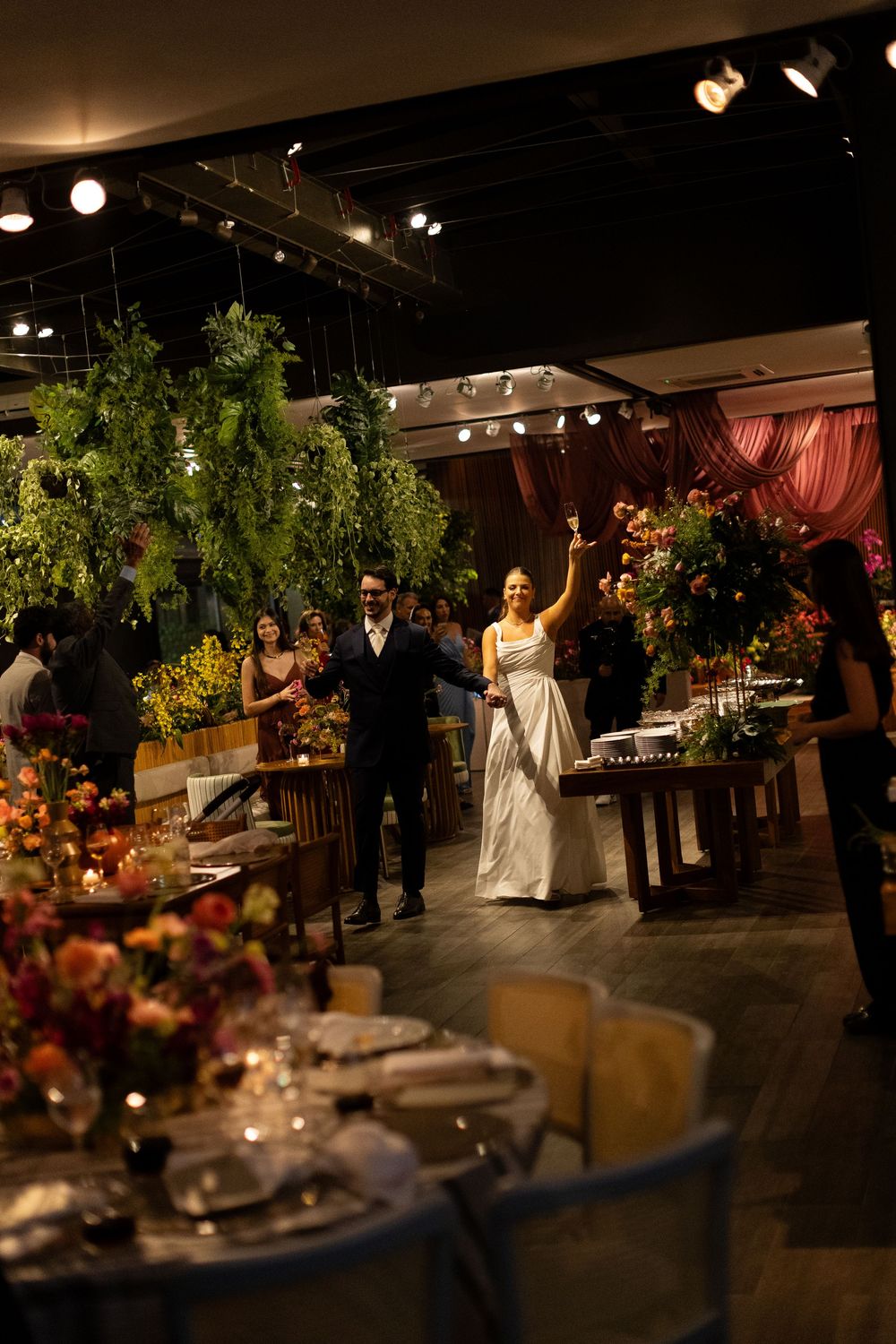Bride and groom make grand entrance at elegant indoor wedding reception with lush floral arrangements and warm ambient lighting.