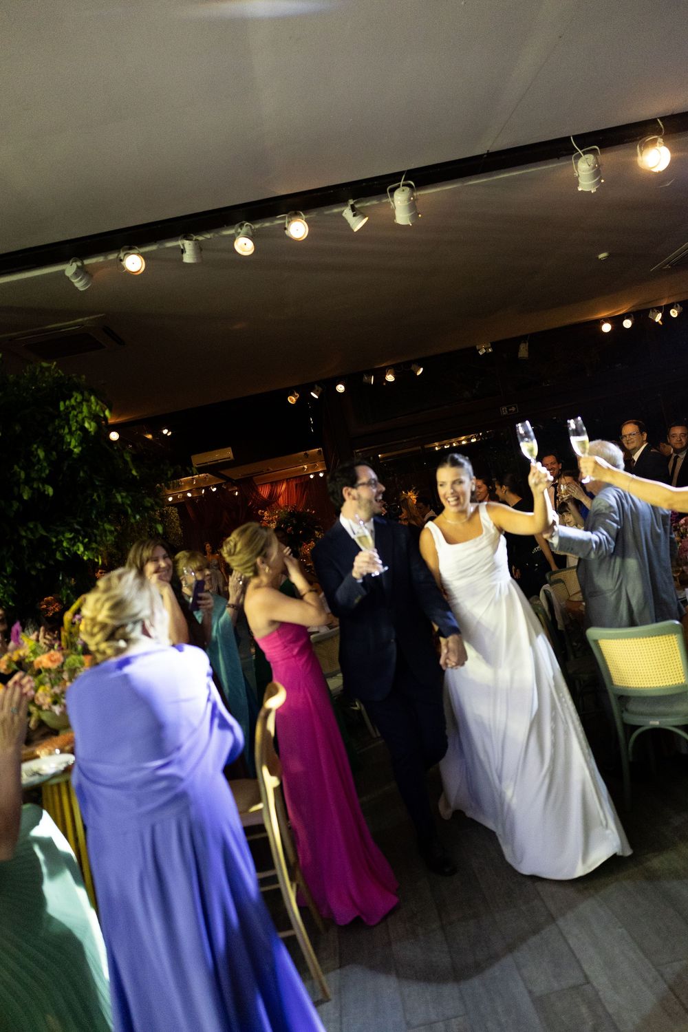 Bride and groom walk through cheering wedding guests raising champagne glasses during indoor reception celebration.