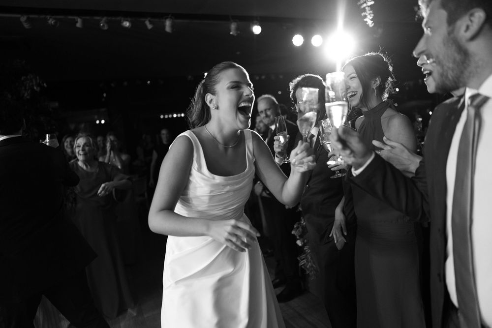 Black and white photo of a joyful bride laughing with guests raising champagne glasses at a wedding reception.