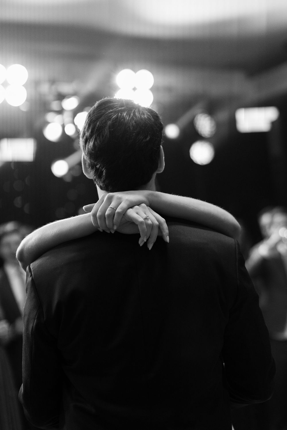 Black and white photo of a couple slow dancing, woman's hand resting on man's shoulder, bokeh lights in background.