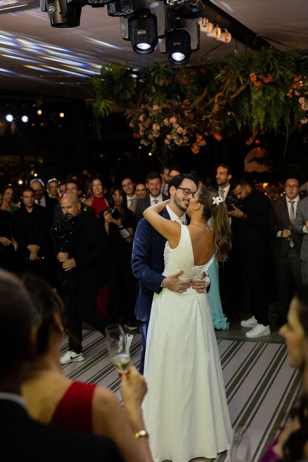 Bride and groom share their first dance at wedding reception surrounded by guests under floral decorations.