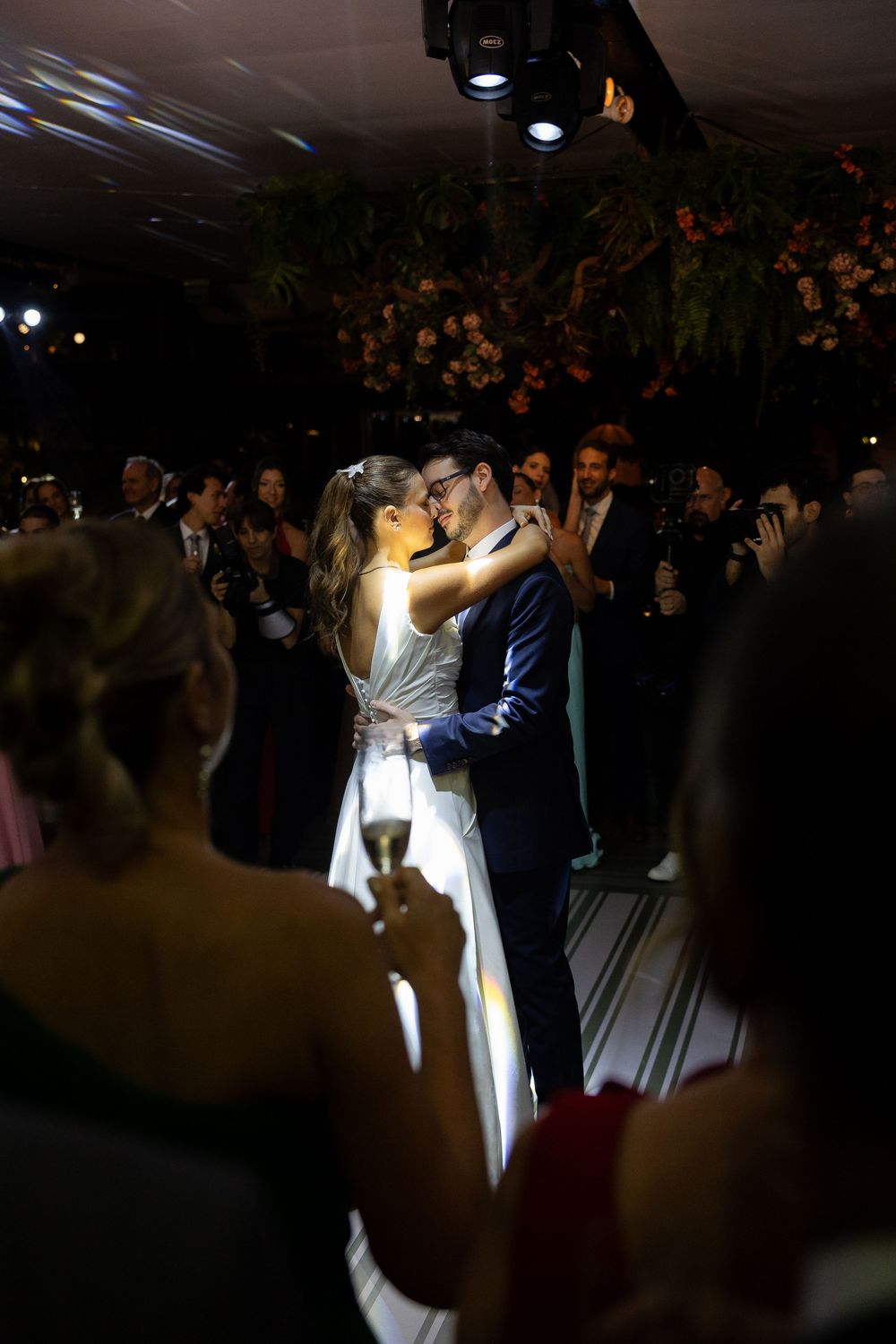 Bride and groom share a romantic first dance kiss surrounded by applauding wedding guests under warm spotlight.