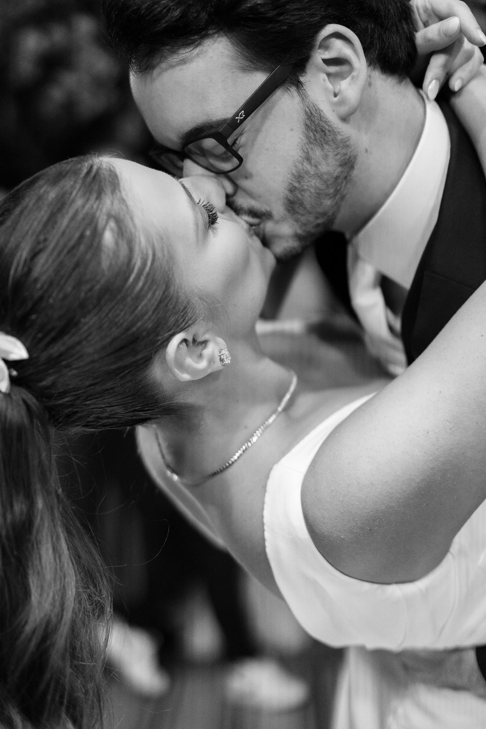 Black and white photo of a couple sharing a romantic kiss, the man wearing glasses and a suit.