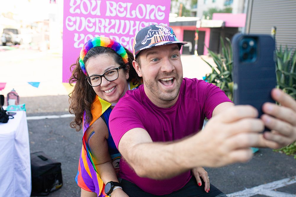Two smiling people take a selfie at a colorful Pride event, with a rainbow outfit and festive backdrop.