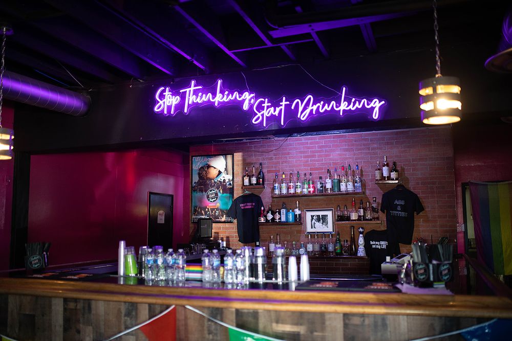 Bar with neon sign reading Stop Thinking Start Drinking, brick walls, liquor bottles, and dark ambiance.