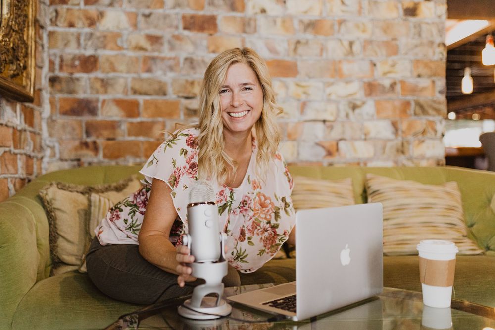 Smiling blonde woman podcasting with a blue Yeti mic and MacBook laptop on a green sofa in a cozy brick-walled cafe.