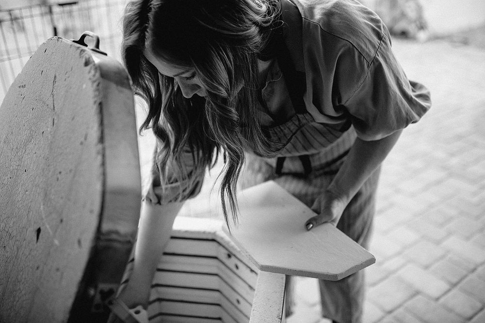 Woman in denim overalls leaning over a wooden stool outdoors in black and white photo.