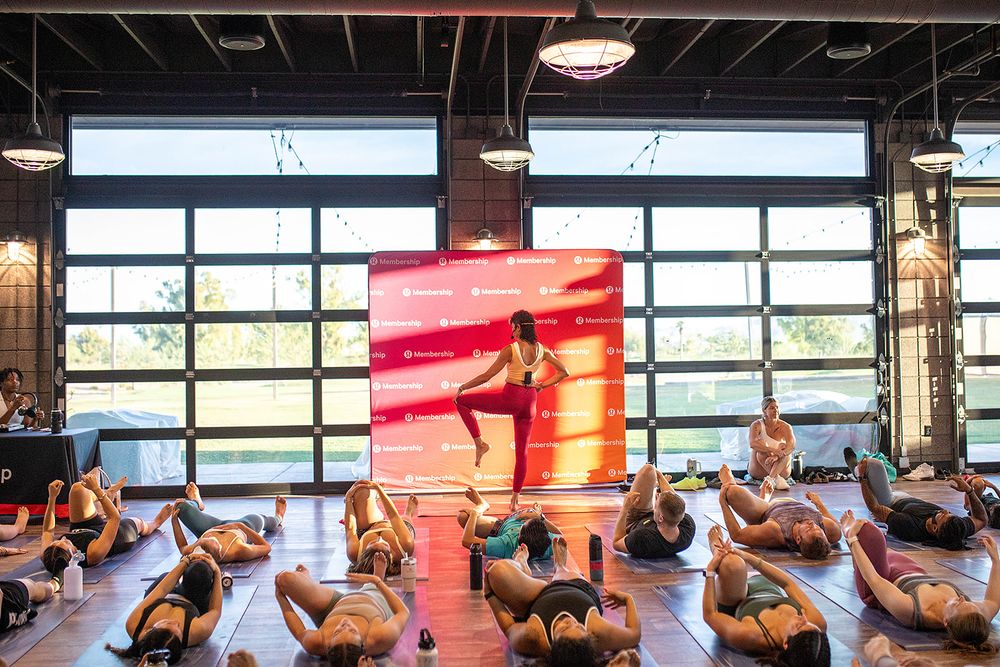 Fitness instructor leading a yoga class with participants lying on mats in a modern studio with large garage-style windows.