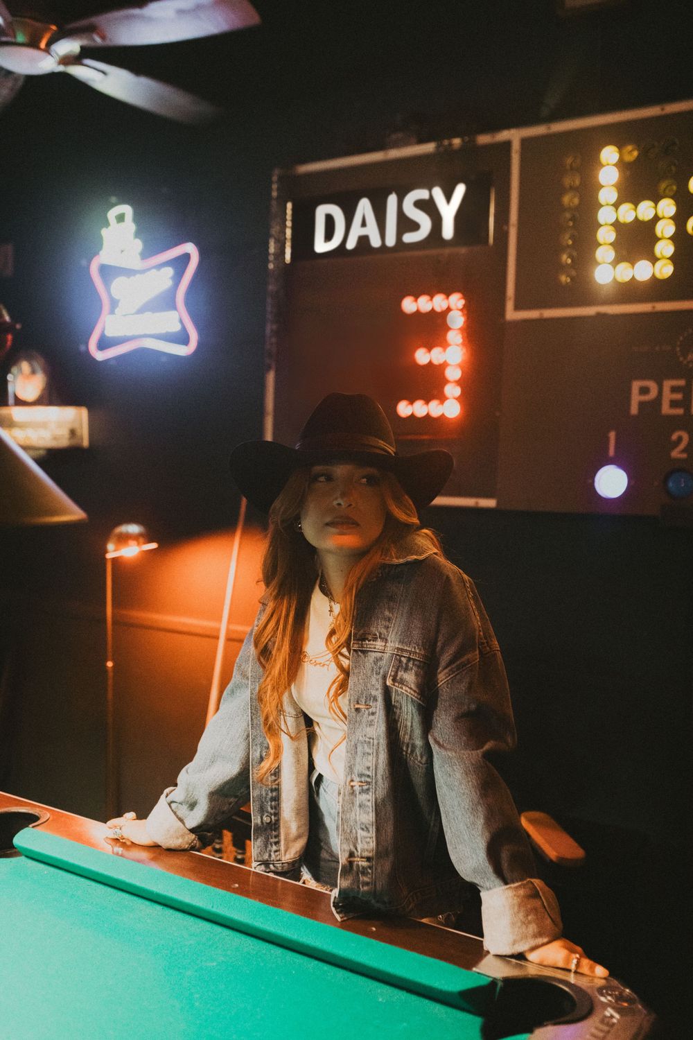 Young woman in denim jacket and cowboy hat playing pool in a dimly lit bar with neon signs.