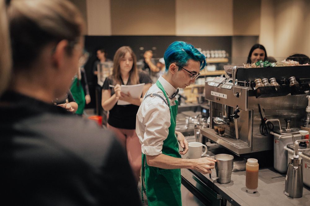 Starbucks barista with teal hair and green apron preparing coffee drinks at an espresso machine in a busy cafe.
