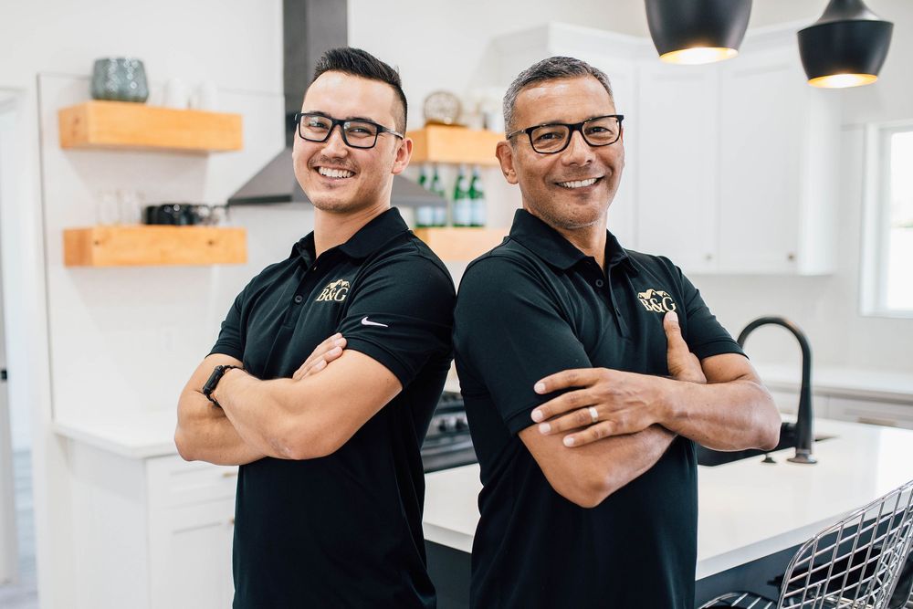 Two smiling men in matching black polo shirts stand back-to-back with arms crossed in a modern white kitchen.
