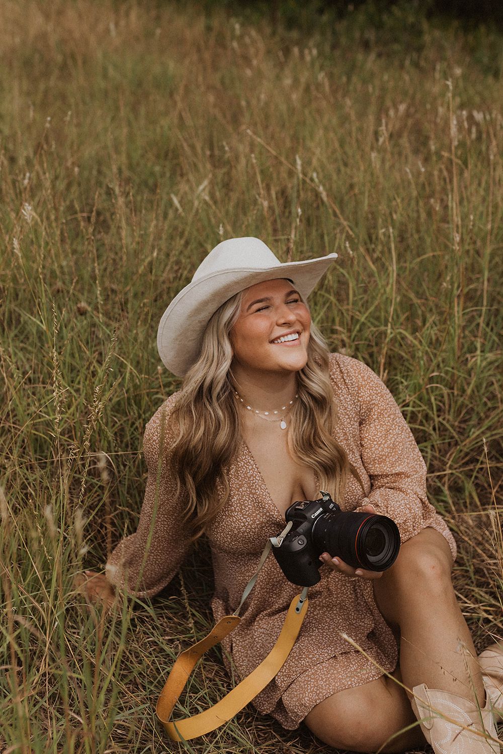 Smiling woman in cowboy hat and floral dress sitting in tall grass holding a DSLR camera with yellow strap.