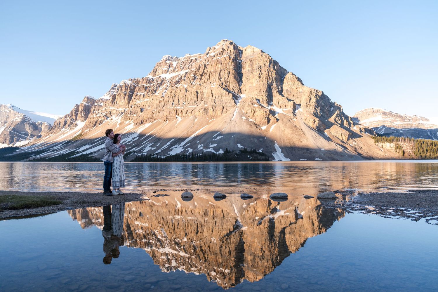 Couple standing by a reflective alpine lake with stunning mountain peaks and snow in Bow Lake, Banff National Park, Canada.