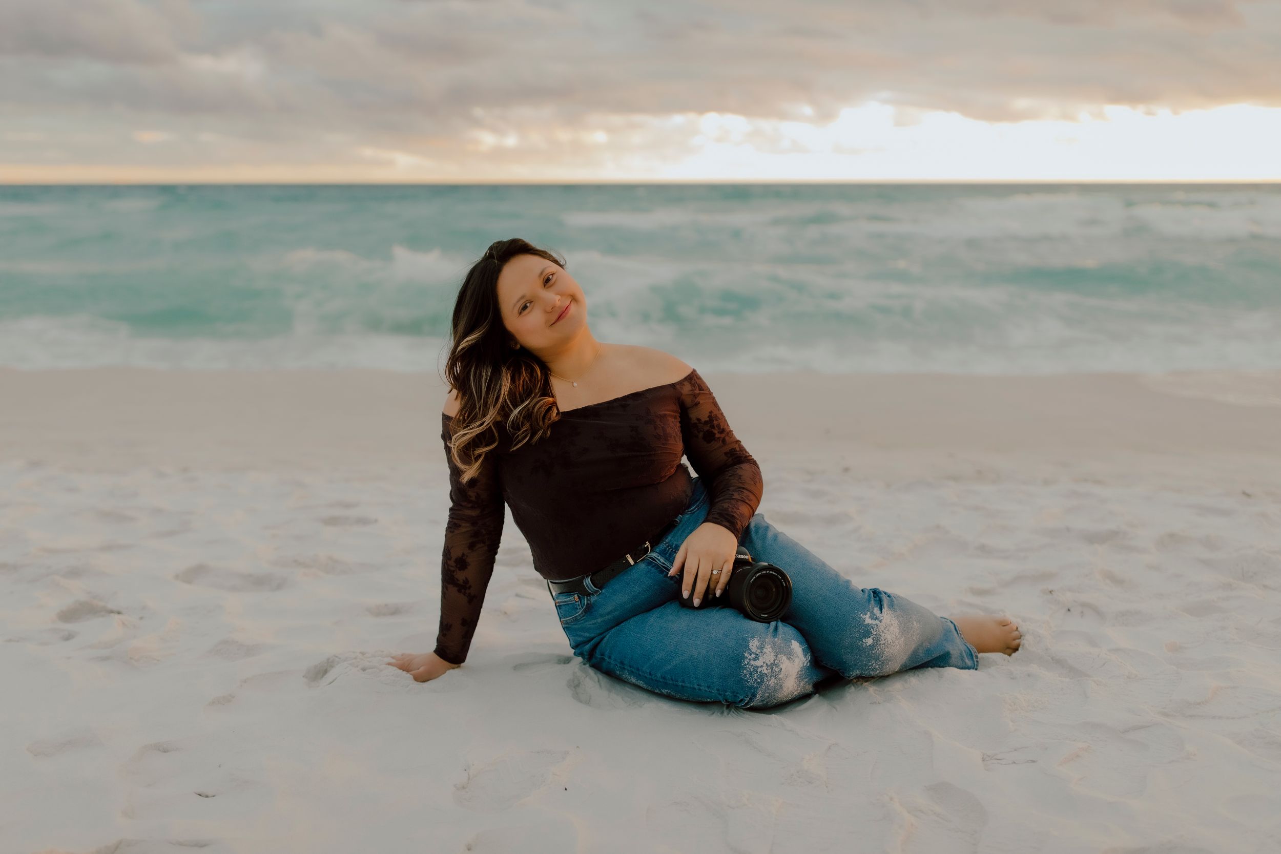 Woman in dark off-shoulder top and ripped jeans sitting on white sand beach at sunset.
