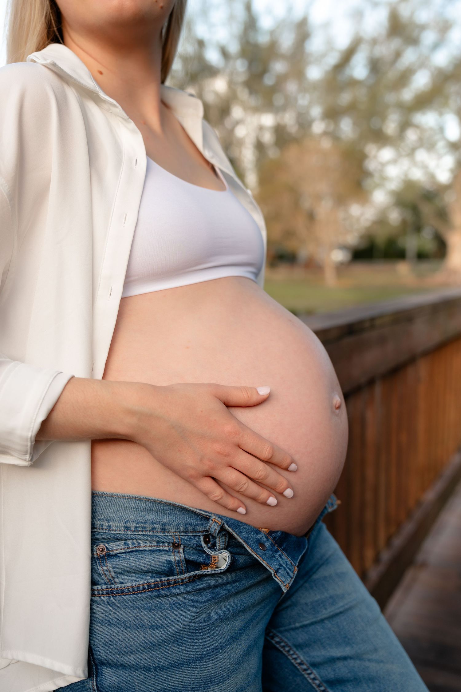 Pregnant woman in white shirt and jeans cradling her bare baby bump outdoors in a park setting.