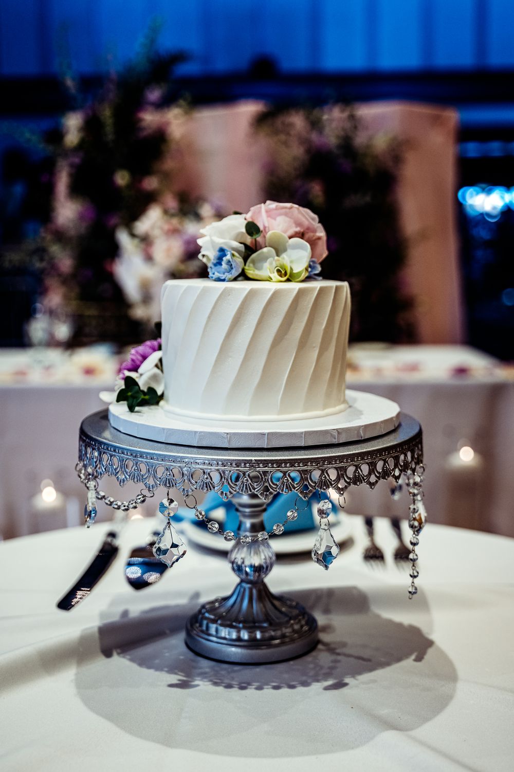 Elegant two-tier white wedding cake with floral topper on ornate silver stand with crystal details.