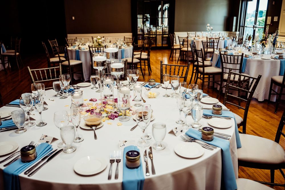 Elegant wedding reception venue with round tables set with white linens, blue napkins, glassware, and floral centerpieces.