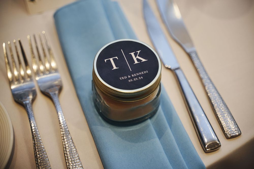 Personalized wedding favor jar with monogram TK on navy label, placed on blue napkin with silverware at elegant reception table.