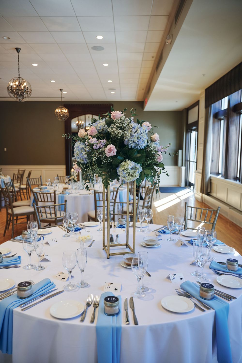 Elegant wedding reception table with blue napkins, crystal glassware, and tall floral centerpiece of pink roses and hydrangeas.